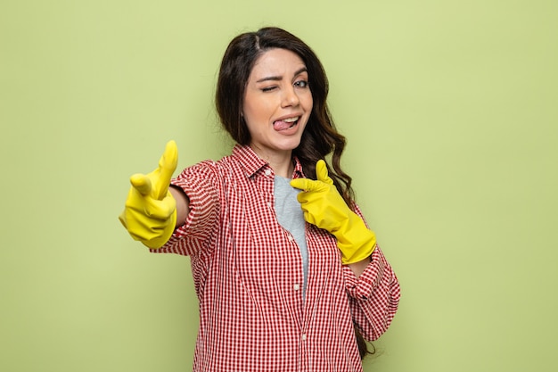 Confident pretty caucasian cleaner woman with rubber gloves sticking out her tongue and pointing at front with two hands