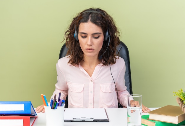 Confident pretty caucasian female call center operator on headphones sitting at desk with office tools looking at clipboard