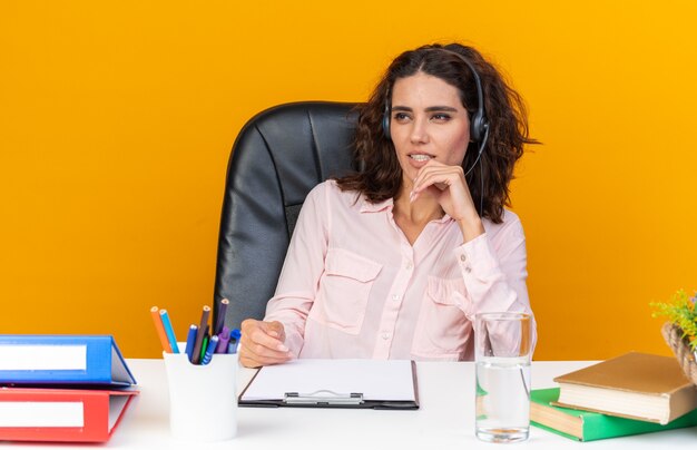 Confident pretty caucasian female call center operator on headphones sitting at desk with office tools looking at side 