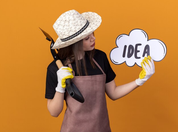 Confident pretty caucasian female gardener wearing gardening hat and gloves holding spade and looking at idea bubble isolated on orange wall with copy space