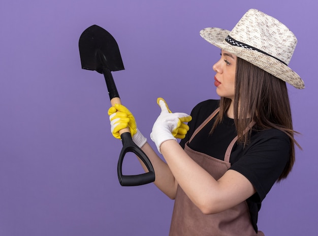 Confident pretty caucasian female gardener wearing gardening hat and gloves looking and pointing at spade