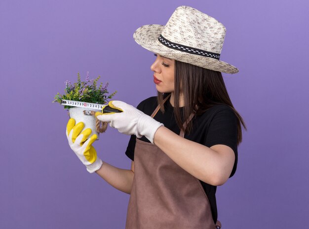 コピースペースで紫色の壁に分離された巻尺で植木鉢の花を測定ガーデニングの帽子と手袋を身に着けている自信を持ってかなり白人女性の庭師