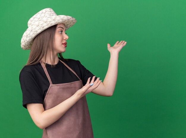 Confident pretty caucasian female gardener wearing gardening hat looking and pointing at side with two hands on green