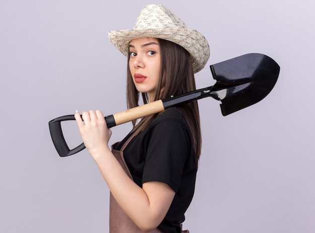 Free photo confident pretty caucasian female gardener wearing gardening hat stands sideways holding spade on shoulder on white