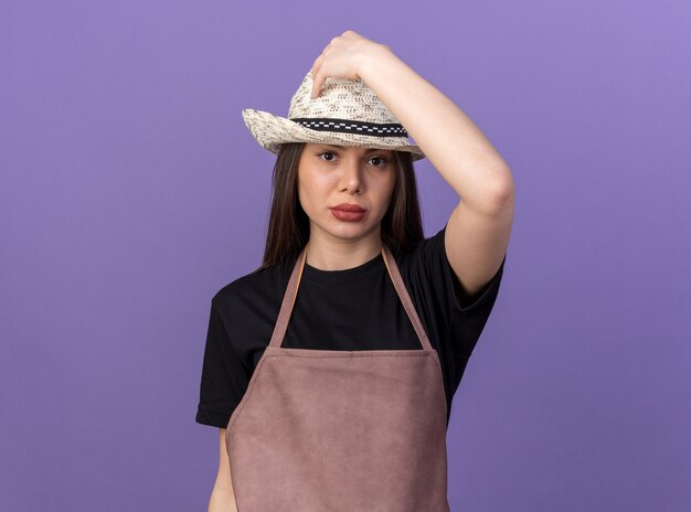 Confident pretty caucasian female gardener wearing and putting hand on gardening hat