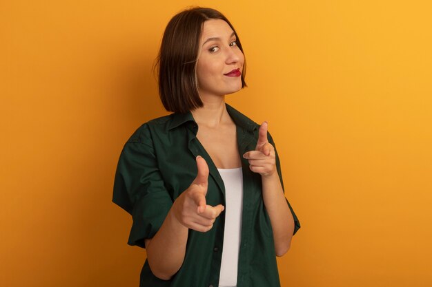 Confident pretty caucasian woman looks and points at camera with two hands on orange