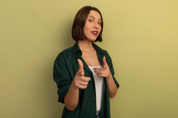 Confident pretty caucasian woman points at camera with two hands on olive green