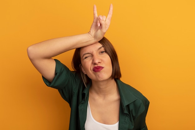 Confident pretty caucasian woman puts hand on head gesturing horns hand sign on orange