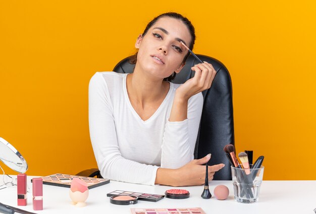 Confident pretty caucasian woman sitting at table with makeup tools applying eyeshadow with makeup brush