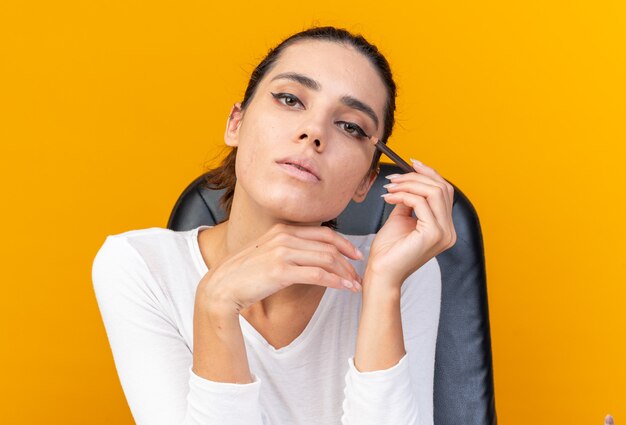 Confident pretty caucasian woman sitting at table with makeup tools holding eyeliner isolated on orange wall with copy space