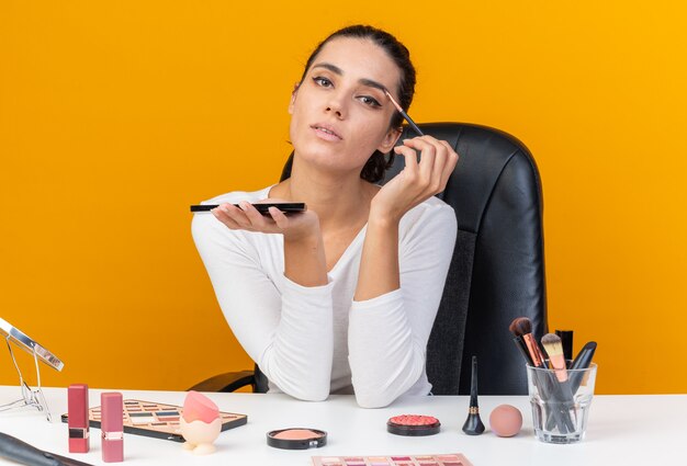 Confident pretty caucasian woman sitting at table with makeup tools holding eyeshadow palette and applying eyeshadow with makeup brush isolated on orange wall with copy space