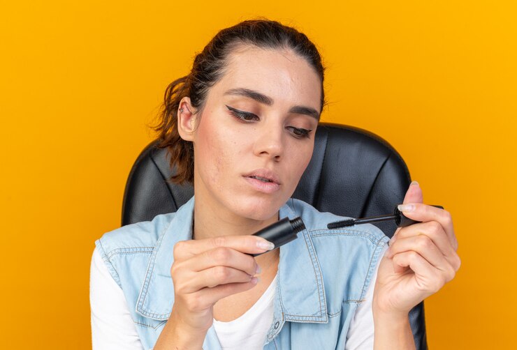 Free Photo confident pretty caucasian woman sitting at table with makeup tools holding and looking at mascara isolated on orange wall with copy space