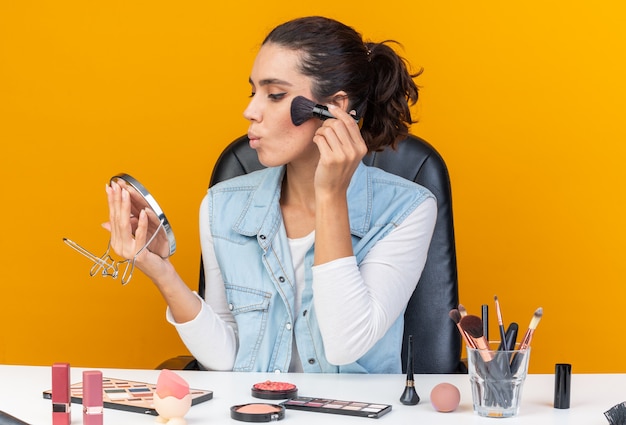 Confident pretty caucasian woman sitting at table with makeup tools holding and looking at mirror applying blush with makeup brush isolated on orange wall with copy space Premium Photo