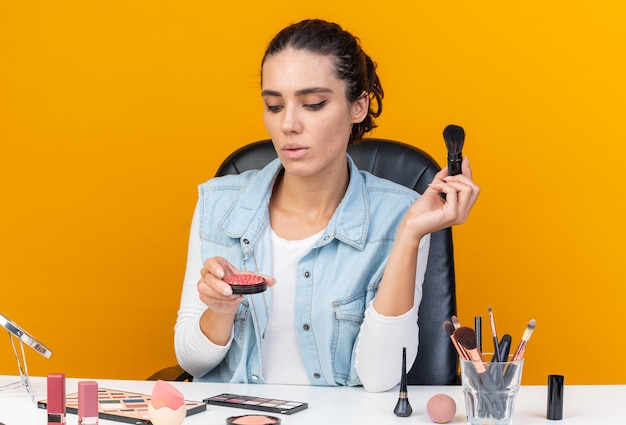 Confident pretty caucasian woman sitting at table with makeup tools holding makeup brush and looking at blush isolated on orange wall with copy space