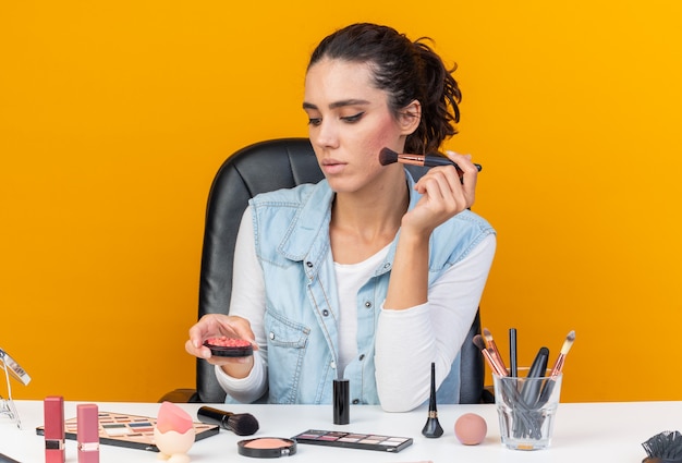 Confident pretty caucasian woman sitting at table with makeup tools holding makeup brush looking at blush 