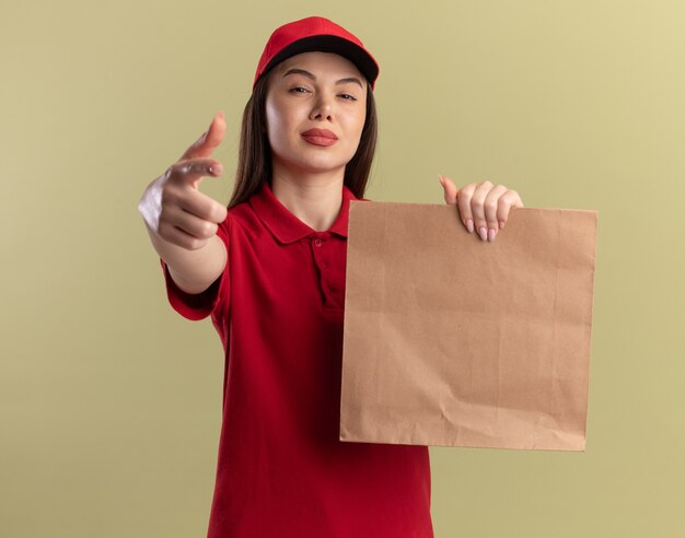 Confident pretty delivery woman in uniform holds paper package and points at camera on olive green