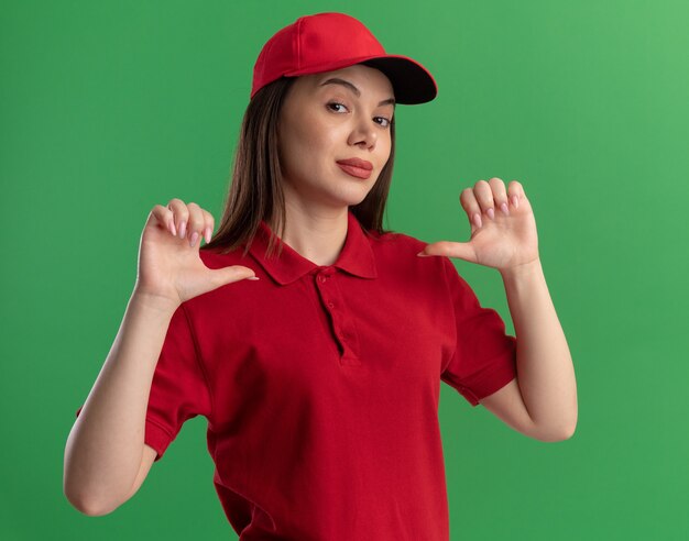 Confident pretty delivery woman in uniform points at herself with two hands on green