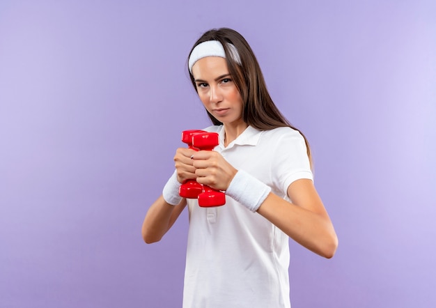Confident pretty sporty girl wearing headband and wristband holding dumbbells isolated on purple wall with copy space