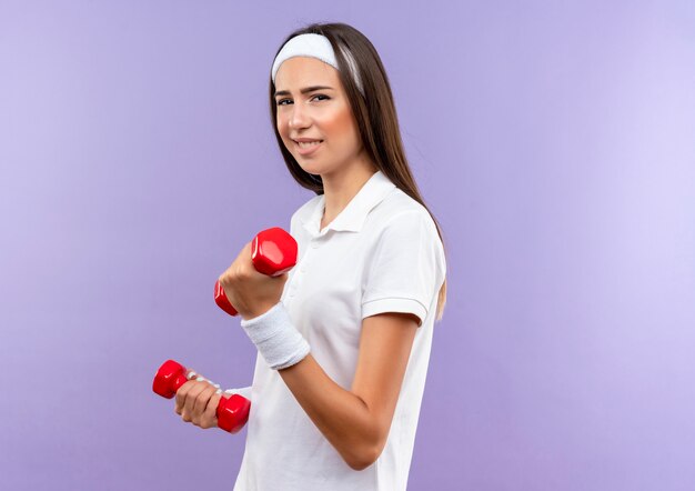 Confident pretty sporty girl wearing headband and wristband holding dumbbells isolated on purple wall with copy space