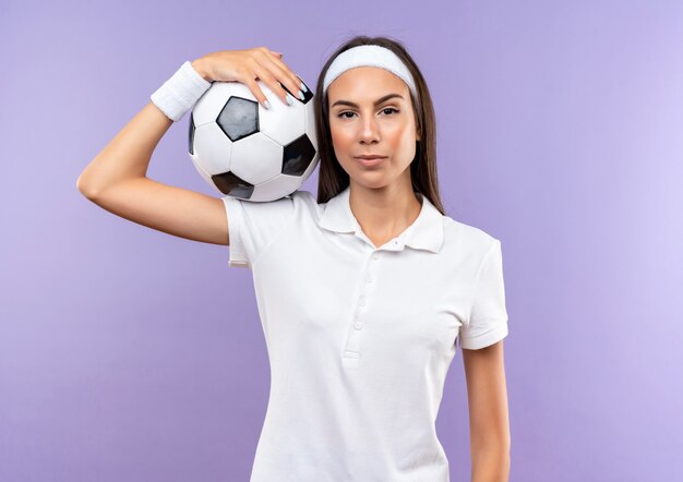 Confident pretty sporty girl wearing headband and wristband holding soccer ball on shoulder isolated on purple wall