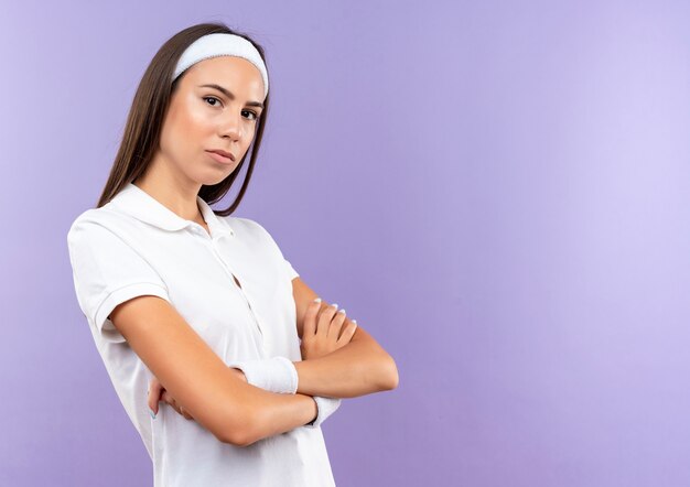 Уверенная в себе симпатичная спортивная девушка с ободком и браслетом стоит в закрытой позе, изолированной на фиолетовой стене с копией пространства