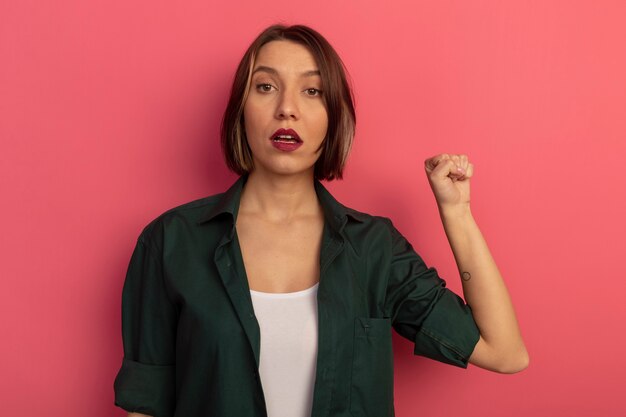 Confident pretty woman keeps fist up isolated on pink wall