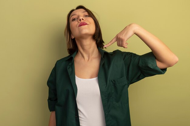 Confident pretty woman points at herself isolated on olive green wall