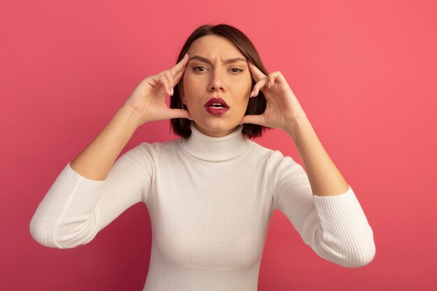 Confident pretty woman puts fingers on temples isolated on pink wall