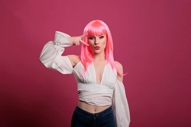 Confident pretty woman with pink hair doing peace sign and kissy face in front of camera. Carefree attractive girl showing victory symbol and peaceful gesture with two fingers up in studio.