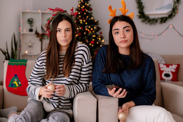 Confident pretty young girls with holly wreath and reindeer headband hold glass ball ornaments sitting on armchairs and enjoying christmas time at home