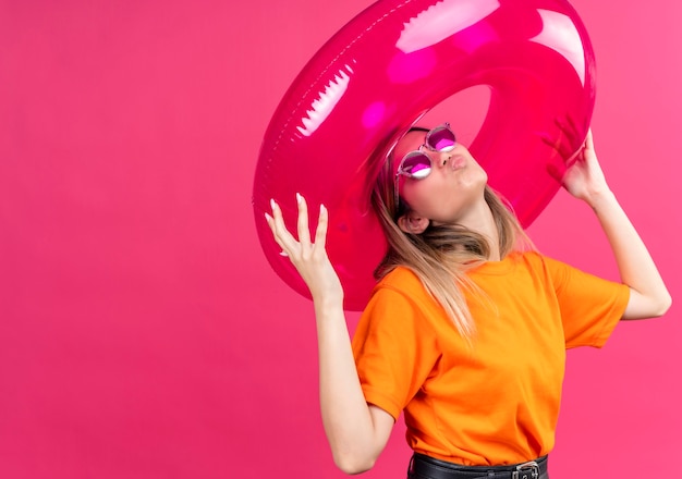 Free photo a confident pretty young woman in an orange t-shirt wearing sunglasses blowing kisses while holding pink inflatable ring on a pink wall