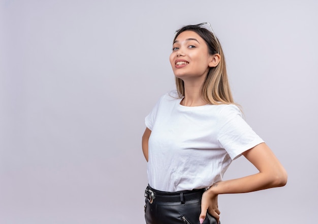 A confident pretty young woman in white t-shirt wearing sunglasses on her head keeping hands on waist while looking on a white wall