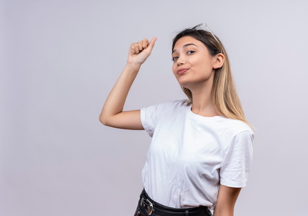 A confident pretty young woman in white t-shirt wearing sunglasses on her head raising clenched fist while looking on a white wall