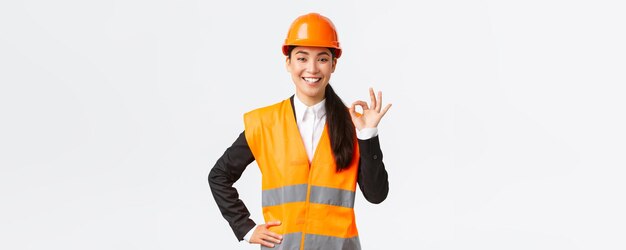 Confident professional asian female architect in safety helmet assure quality and work in time showing okay gesture and smiling determined standing assertive ensure and guarantee something