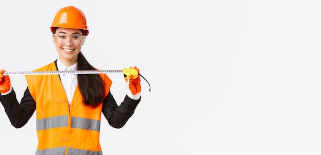 Free photo confident professional female asian architect measuring layout wearing safety helmet and uniform and holding tape measure smiling pleased satisfied with achieved result during construction