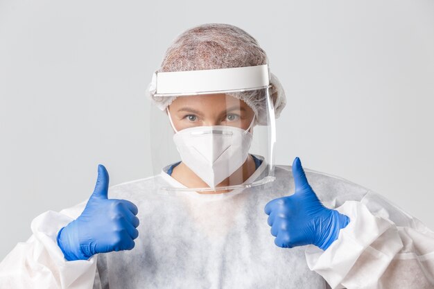 Free photo confident professional female doctor in personal protective equipment, face shield and respirator showing thumbs-up, saving patient lives