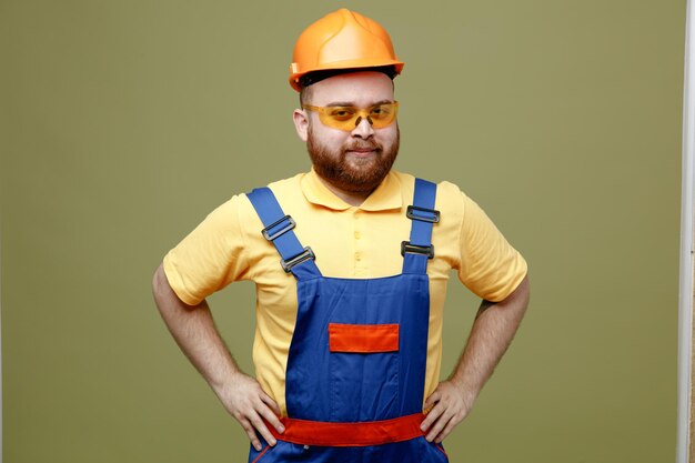 Confident putting hands on hips young builder man in uniform isolated on green background