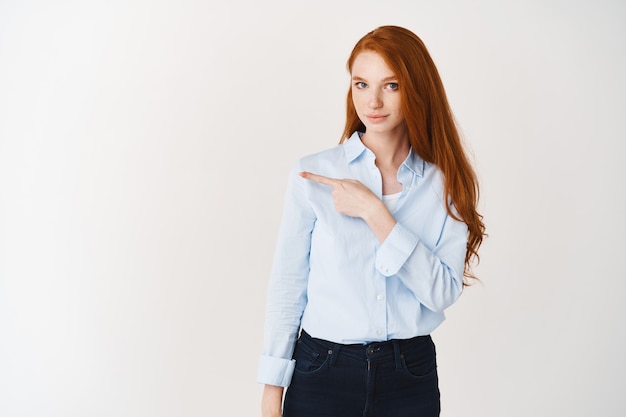 Confident redhead businesswoman pointing finger left, showing company logo on white wall, wearing blue shirt