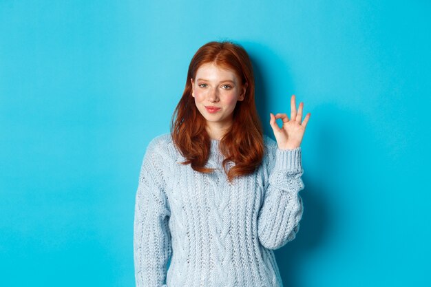 Confident redhead girl assuring you, showing okay sign and smiling, saying yes, approve and agree, standing over blue background