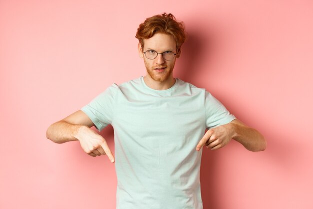 Уверенный рыжий мужчина в очках, указывая пальцами вниз, дерзко глядя с самодовольным лицом в камеру, показывая рекламу, стоя на розовом фоне.