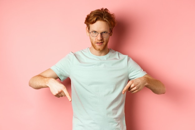 Confident redhead man in glasses pointing fingers down, staring daring with smug face at camera, showing advertisement, standing over pink background.