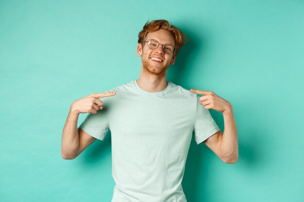 안경과 티셔츠를 입은 자신감 있는 빨간 머리 남자, 늠름한 얼굴로 웃고 자신을 가리키며 청록색 배경 위에 서 있는 동안 자랑합니다.