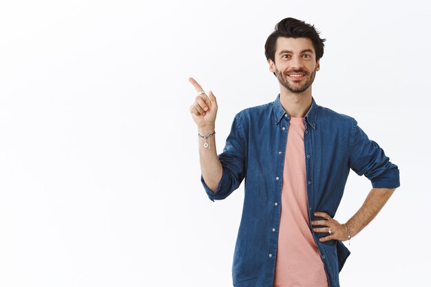 Confident, sassy young bearded gay man in pink t-shirt, shirt, hold hand on waist, pointing upper left corner and smiling, suggest friends visit party nearby