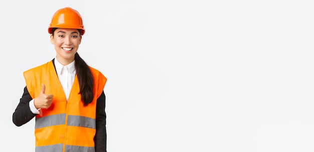 Confident satisfied asian female construction manager in safety helmet reflective jacket showing thumbsup in approval support and like work of employees standing white background