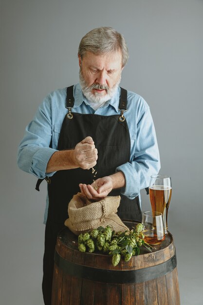 Confident senior man brewer with self crafted beer in glass on wooden barrel on grey wall. Owner of factory presented his products, testing quality. 