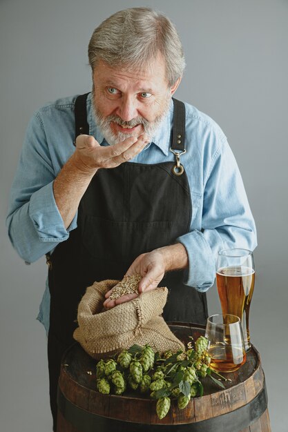 Confident senior man brewer with self crafted beer in glass on wooden barrel on grey wall