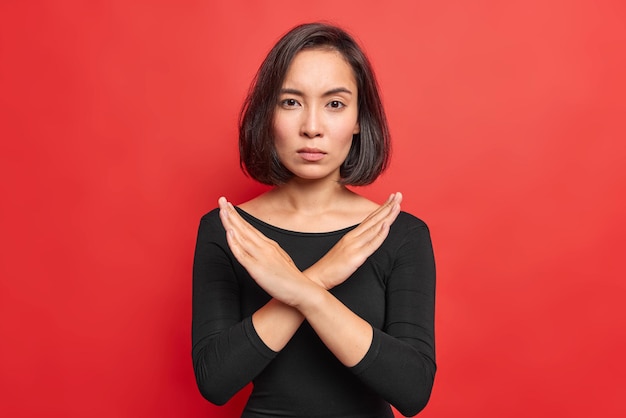 Confident serious Asian woman crosses hands makes stop forbid sign rejects something and says no wears black jumper isolated over vivid red background says no has determined face expression.