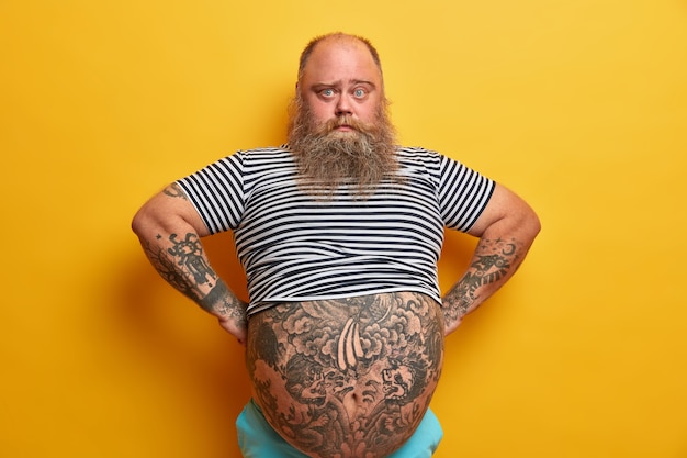 Confident serious blue eyed man with beard, has big abdomen, leads unhealthy lifestyle, dressed in striped undersized sailor t shirt, poses over yellow wall. plump guy stands self assured indoor