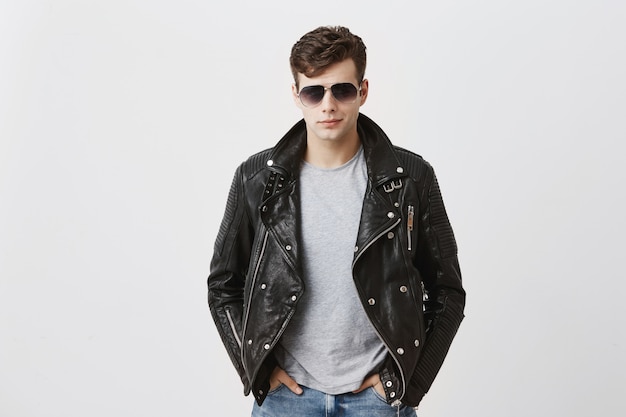 Confident serious handsome man wears black leather jacket over gray t-shirt and stylish eyewear, looks directly into camera, isolated . people and style concept