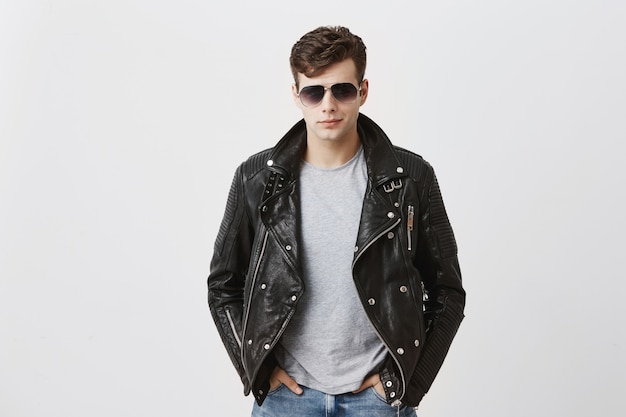 Confident serious handsome man wears black leather jacket over gray t-shirt and stylish eyewear, looks directly into camera, isolated . People and style concept